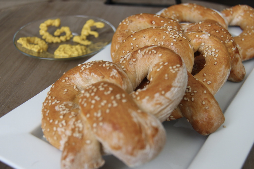 A number of pretzels. In the background, a small, clear plate is covered with mustard which spells OGE.