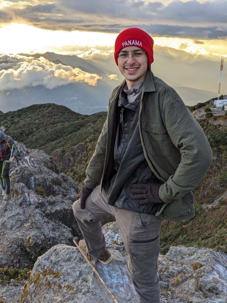 John stands on top of a mountain. He wears an olive-green jacket and a bright red beanie with the word “Panama” in white text on the front. 
