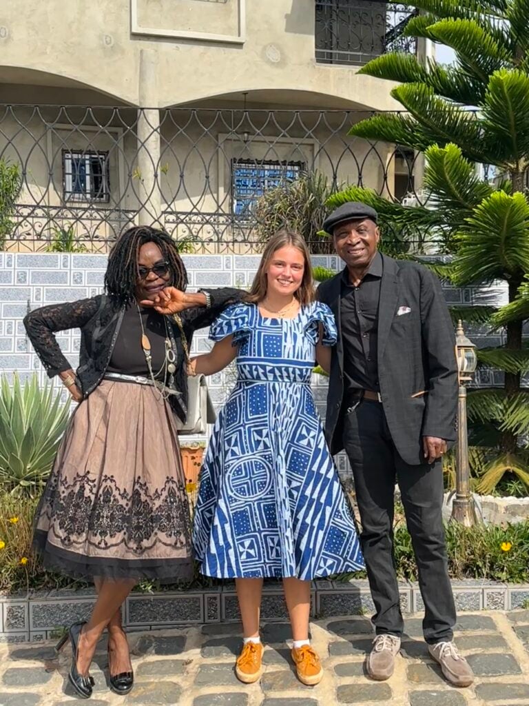 A girl in a blue dress in yellow shoes stands in between her host parents. The mom wears a black outfit with a brown patterned skirt on the left, and the father wears a black suit
