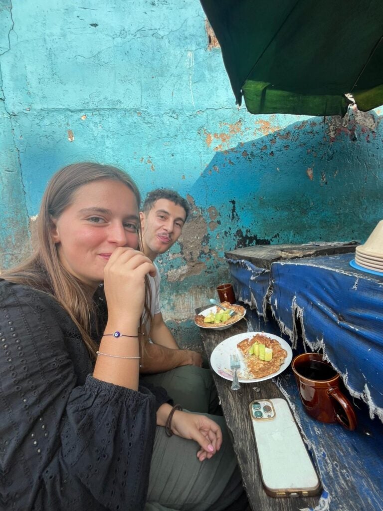 A girl with blonde hair and a boy with dark curly hair sits behind her against a bright blue wall. They are eating a spaghetti omelette and drinking tea